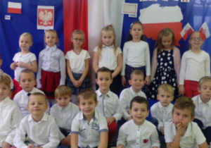 Cała grupa dzieci ubrana w stroje galowe pozuje do zdjęcia na tle dekoracji przedstawiającej symbole narodowe.
